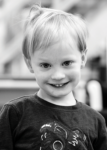 A black and white photo of Fitz, a student at Rosen Family Preschool. They have short blonde hair and light skin. They are looking at the camera and smiling.